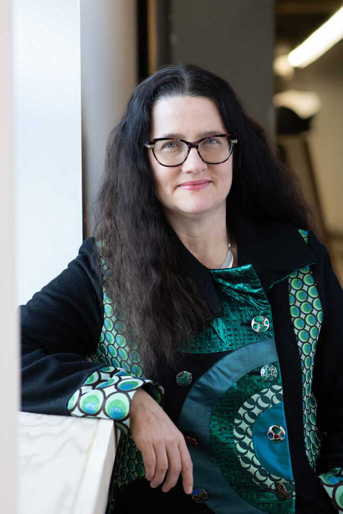 A female-bodied person with dark wavy hair and glasses in a brilliant green coat.